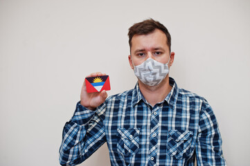 Man in checkered shirt show Antigua and Barbuda flag card in hand, wear protect mask isolated on...
