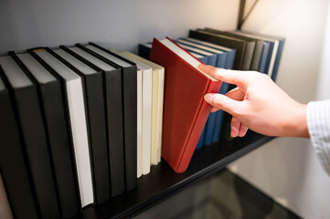 Male hand choosing and picking red book from white bookshelf in public library. Education research...