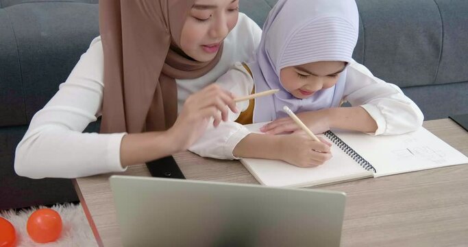 Beautiful young traditional Islamic muslim Asian mother in house life style working teach her daughter education homework from laptop PC on the wooden table and writing to book