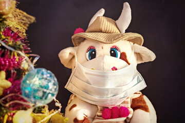 White soft toy bull in a medical mask on a dark background with a Christmas tree and garlands, New Year's card. New Year and Christmas during the coronavirus pandemic