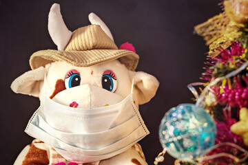 White soft toy bull in a medical mask on a dark background with a Christmas tree and garlands, New Year's card. New Year and Christmas during the coronavirus pandemic