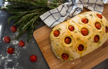 Fresh baked homemade focaccia bread with rosemary and cherry tomatoes on grey textured background. Top view photo. Traditional cooking concept. 