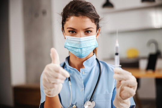 Portrait Of Nurse With Face Mask On Sitting At Home, Holding Syringe With Vaccine And Showing Thumbs Up During Corona Virus.