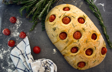 Fresh baked homemade focaccia bread with rosemary and cherry tomatoes on grey textured background. Top view photo. Traditional cooking concept. 