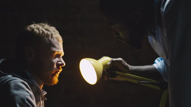 Police officer pressing and interrogating man suspected of crime glowing light of lamp into face.