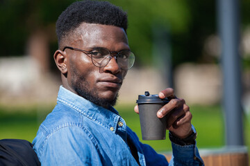 Close up of young African-American businessman in stylish clothes sitting at bench in a city park and drinking hot coffee