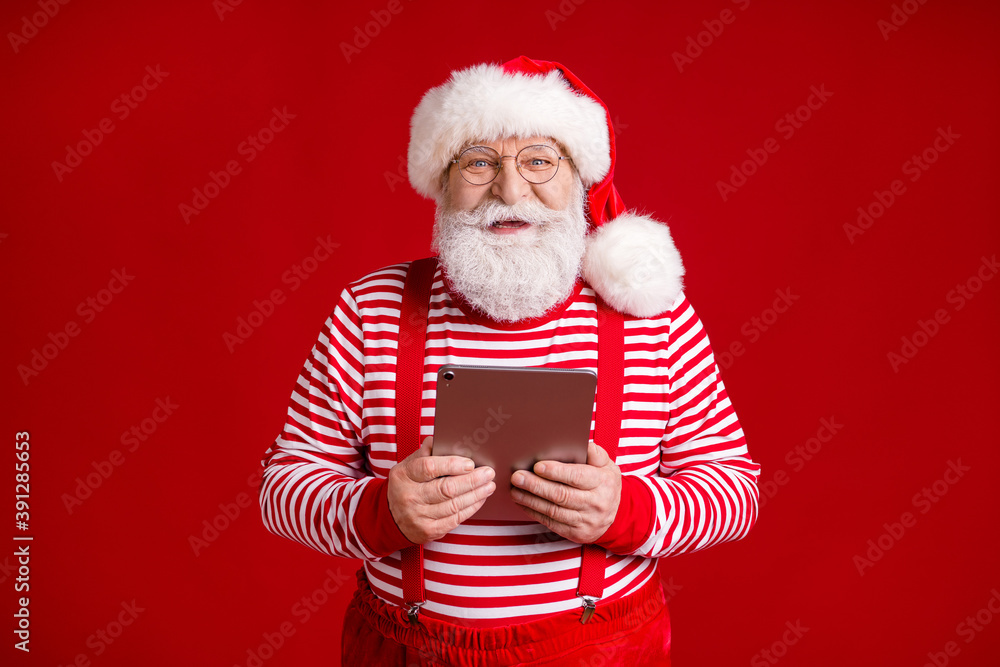 Wall mural Photo of pensioner old man grey beard hold tablet search fastest sledge way north pole wear santa x-mas costume suspenders spectacles striped shirt cap isolated red color background