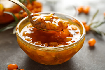 Spoon with delicious sea buckthorn jam over bowl on grey table, closeup