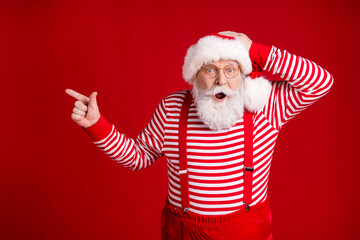 Fototapeta na wymiar Photo of retired old man grey beard direct finger empty space hand head shocking baubles sale wear santa x-mas costume suspenders spectacles striped shirt cap isolated red color background