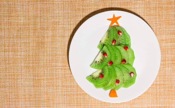 Christmas edible tree made from kiwi, carrot and pomegranate on plate on the table. Dessert idea for kids. New Year food art concept. Top view, copy space