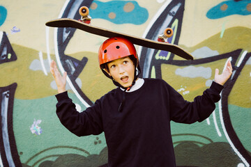 Portrait of a young skater boy wearing  red helmet and playing with an old skateboard, in front of a graffiti mural