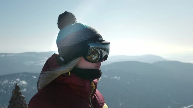 Portrait of a beautiful girl looking at the mountainside. A tourist in a mask or snowboard goggles looks ahead. The concept of victory in sports. Winter travel at the ski resort.