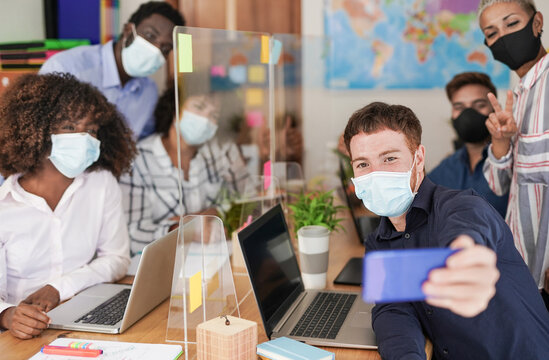 Young multiracial people in office taking a selfie while wearing protective face mask - Work places with safety measures