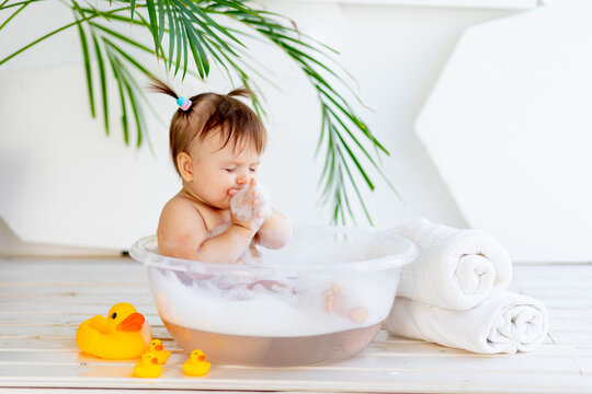 funny little baby girl washes in a basin with foam and water in a bright room at home