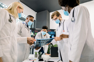 Fototapeta premium Covid outbreak concept. Computed tomography. Group of multiracial doctors radiologists in facial masks brainstorming and discussing the diagnosis while watching patient's lungs CT scan