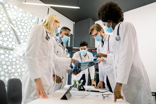 Team of diverse confident multiethnic doctors in masks, holding and looking at patient CT scan, discussing diagnosis and treatment. CT scan, radiology concept. Computed tomography and covid