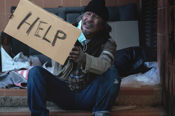 Homeless begging man with a medical mask sits on the steps holding a brown cardboard, The word "Help" is on the cardboard