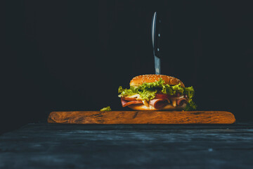 Homemade burger with ham, cheese, salad and pomegranate, pierced by a knife on a wooden board