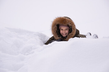 A smiling little girl in a winter jacket with a fur hood lies in a large snowdrift in a white field