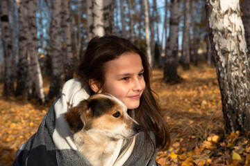 A smiling girl with long hair hugs a dog in a birch grove against the background of fallen yellow leaves