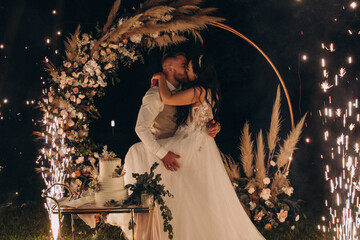 The bride and groom congratulate each other against the background of a flower arch. Evening wedding. Fireworks and lights.