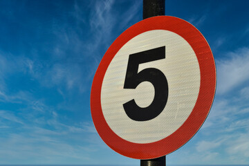5 miles per hour road sign UK. Circular with red outline, white background and black number five. Isolated against blue sky and light clouds background.