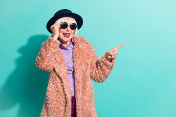 Photo of cute old woman dressed vintage outerwear cap eyewear pointing empty space isolated...