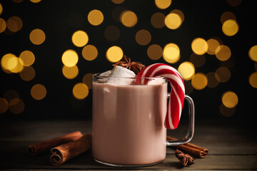 Obraz premium Glass cup of tasty cocoa with marshmallows and Christmas candy cane on wooden table against blurred festive lights