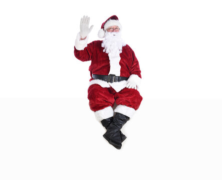 Senior man in traditional Santa Claus Suit sitting on a white wall waving.  Isolated on white with copy space.
