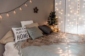 Decorated room for Christmas holidays with bed with a dark blanket, pillows, candles and glowing garland. 