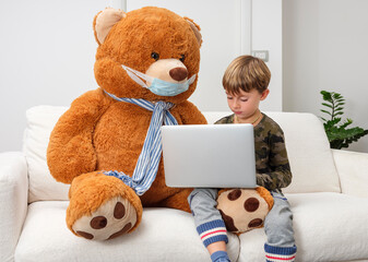 Child takes lessons from home with his laptop, sitting on the sofa with his teddy bear wearing the...