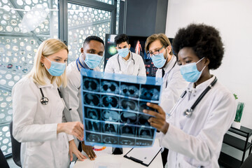 Team of multiethnic doctors or scientists, wearing protective medical face masks, looking at the CT scan of the patient and discussing the results of new treatment or clinical trial. Focus on faces