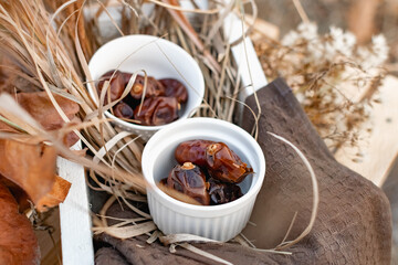 Dried dates, sweet fruits of the date palm in small ceramic forms in a wooden white box, decorated with dry leaves and yellow grass, various autumn twigs. Composition on a stool, late autumn