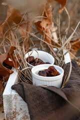 Dried dates, sweet fruits of the date palm in small ceramic forms in a wooden white box, decorated with dry leaves and yellow grass, various autumn twigs. Composition on a stool, late autumn