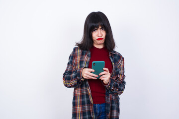 Portrait of a confused Beautiful young Caucasian woman standing against white background holding mobile phone and shrugging shoulders and frowning face.