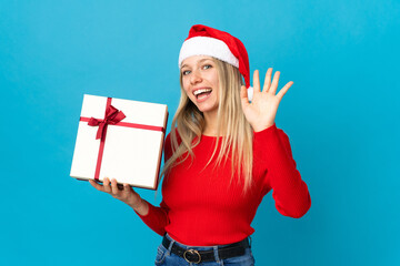 Woman with christmas hat holding a present isolated on blue background saluting with hand with happy expression