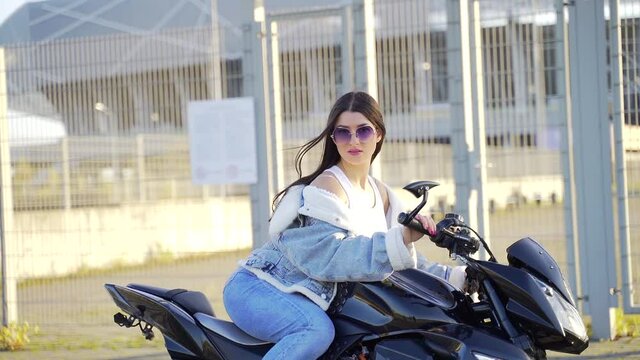 Portrait of an attractive young adult stylish girl sitting on a sports motorcycle. Sexy fashion woman in denim clothes and sunglasses with a bike on urban city background. Looking at camera and posing