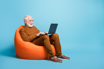 Portrait of his he nice attractive focused bearded grey-haired man sitting in bag chair using laptop writing email letter browsing web wi-fi isolated over blue pastel color background