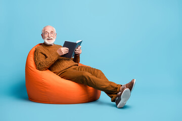 Portrait of his he nice attractive cheerful cheery glad bearded grey-haired man sitting in bag chair reading interesting book enjoying weekend isolated over blue pastel color background