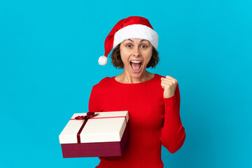 English girl with christmas hat holding a present isolated on blue background celebrating a victory in winner position