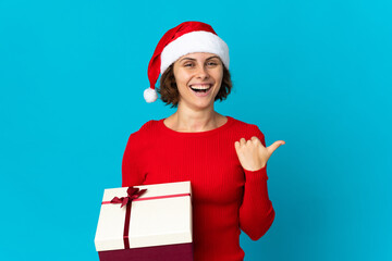 English girl with christmas hat holding a present isolated on blue background pointing to the side to present a product