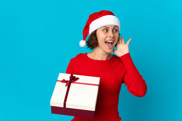 English girl with christmas hat holding a present isolated on blue background listening to something by putting hand on the ear