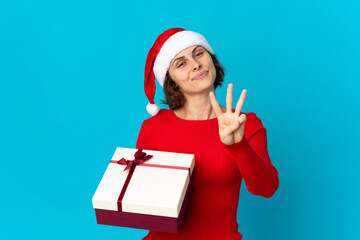 English girl with christmas hat holding a present isolated on blue background happy and counting three with fingers