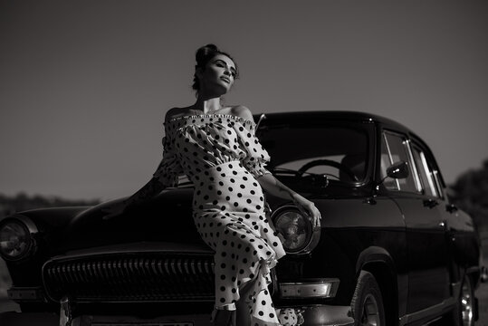 Gorgeous young woman wearing classy spot print off shoulders dress standing on the road leaning on her vintage car