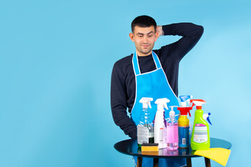 Gender stereotypes. The man holds his head and looks at the set of cleaning products. Photo on a blue background with empty side space.
