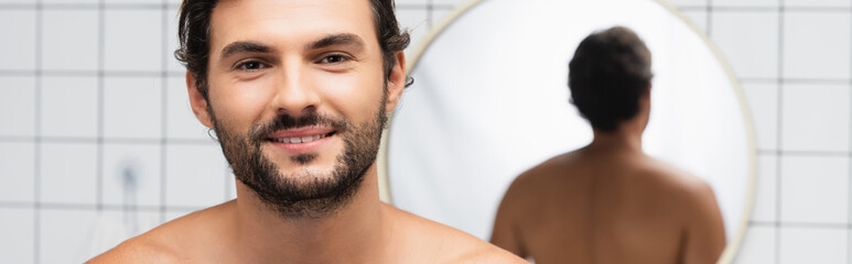 Obraz premium Bearded and shirtless man smiling at camera near mirror on blurred background in bathroom, banner