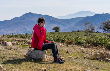 Naklejka na ściany i meble Young woman in red mountain clothes, talking on her cell phone and contemplating the valley