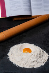 Preparation for a Cookie dough. A pile of flour with an egg in the middle on a slate plate. A rolling pin and a Book with Recipes in the background.