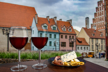 Two glasses of wine with meat and cheese snacks with view of square with the flowers in the old Riga, Latvia. Glass of red wine with different snacks - plate with ham, sliced, blue cheese.