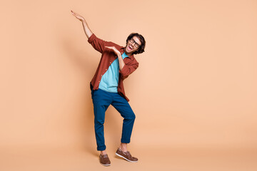Fototapeta na wymiar Full length photo of guy dance raise hands have fun wear spectacles brown t-shirt trousers shoes isolated beige color background